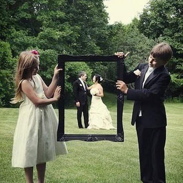 Мальчик и девочка держат рамку картины на природе, а за ней невеста и жених   