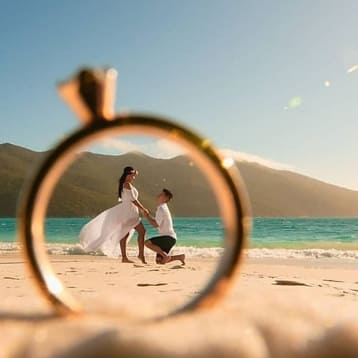 На песке лежит кольцо, а за ним мы видим жениха и невесту радостные на пляже 
